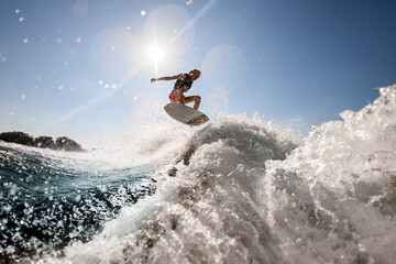 man professionally rides and bounces on surf style wakeboard over splashing wave against blue sky