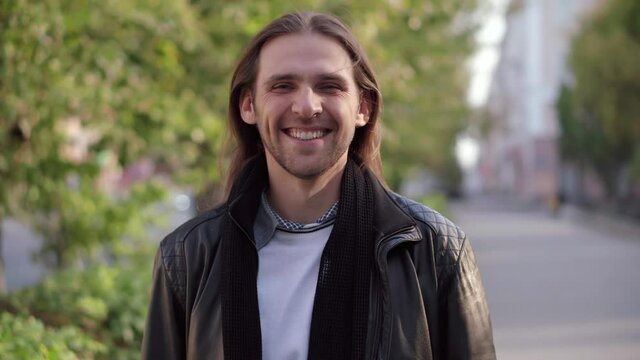 Smiling bearded long-haired man looking at camera. Portrait of a happy handsome young man in a urban street.