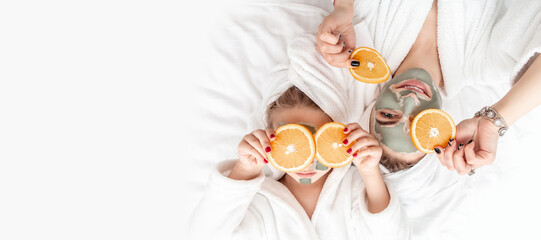 Skin care, beauty, spa. mother and daughter in white bathrobes applying pieces of orange to their eyes, in a white bath towel, posing to camera at home, touching face