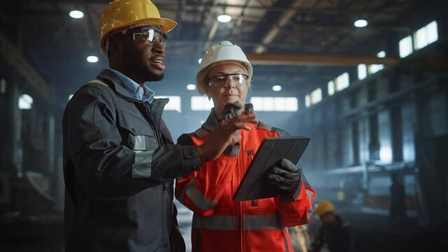 Two Heavy Industry Engineers Stand in Steel Metal Manufacturing Factory, Use Digital Tablet Computer and Have a Discussion. Black African American Industrial Specialist Talk to Female Technician.