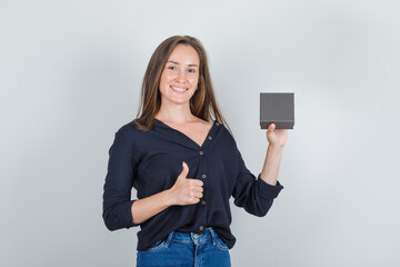 Naklejka premium Young woman in black shirt, jeans shorts holding watch box with thumb up and looking glad , front view.