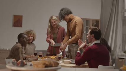 Medium shot of young mixed-race man with knife and fork in his hands standing at dining table and cutting roasted turkey while other people waiting for him and chatting