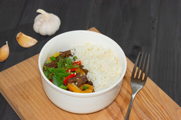 Roast beef with sweet pepper and white rice. White plate, garlic in the background, dark background.