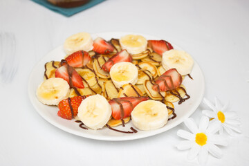 Trendy food - pancake cereal. Heap of mini cereal pancakes on a white plate on a white background. Strawberry slices on a plate, drizzled with syrup. Camomile flowers nearby.