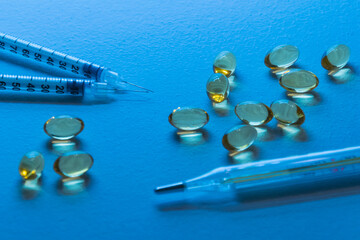 On a blue background background are two syringes and a group of yellow capsules. In the foreground is a thermometer and several capsules. The foreground is blurred.