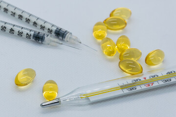 On a white background is a thermometer, next to a pair of yellow plates. In the background are two syringes and a group of yellow capsules. The background is blurry.