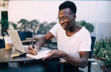 Skilled dark skinned male blogger in trendy wear planning online project making notes in notepad,...