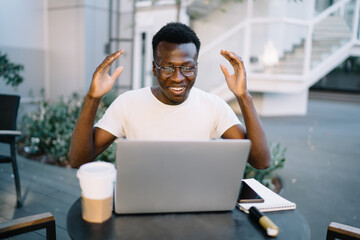 Happy emotional dark skinned male freelancer sitting on cafe terrace overjoyed with getting payment from online successful project, young african american man watching video celebrating victory