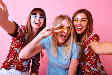 Happy three smiling stylish girls having fun , showing peace gesture and smiling, trendy hipster sunglasses and fashion color matching clothes, friendship goals, pink background.