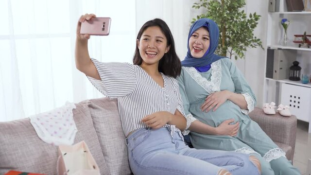 happy girl putting up the peace sign and touching her face while using the phone to take a selfie photo with her Islamic pregnant friend in cozy bright living room.