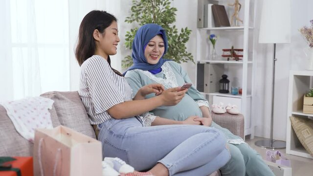 muslim woman in her last trimester is having a hearty laugh with her young female companion while watching fun pictures and videos on phone at home.