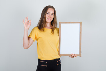 Young woman holding white board with ok sign in t-shirt, pants and looking glad. front view.