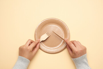 empty white paper plate and female hands are holding disposable fork and knife on beige background