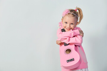 Cute caucasian little girl 4-5 year old in pink sweatshirt are playing the ukulele against gray studio background