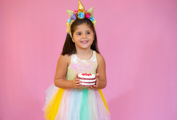 A little girl with dark hair holds a birthday cake on a pink background