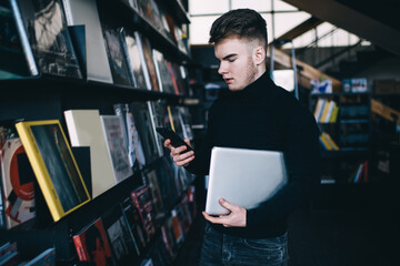 Man with laptop using social media on smartphone in library