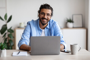 Happy Indian Freelancer Man Sitting At Desk With Laptop, Smiling At Camera