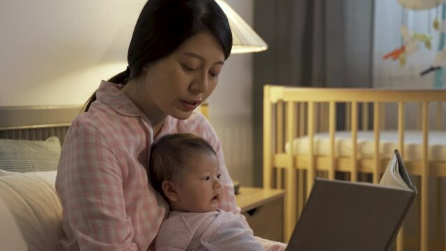 cropped shot Chinese mom is pointing to the book while reading a bedtime story to her baby girl in the bedroom at night.