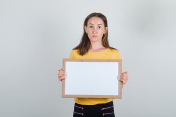 Fototapeta na wymiar Young woman holding white board in yellow t-shirt, black pants and looking careful. front view.