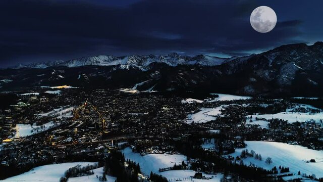 Mountains at winter night, aerial view. Magical Christmas village landscape. Illuminated alpine city covered with snow, white mountains, ski resorts and big shining moon. Zakopane