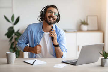 Cheerful indian freelancer guy singing and listening music with headphones while working