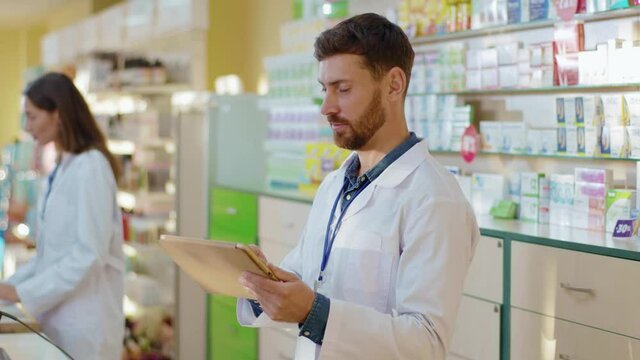 Male caucasian pharmacy intern exploring medications with digital tablet asking advice from professional apothecarist cooperating in drugstore. Healthcare and medicine.