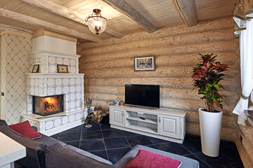 Rustic style living room with fireplace and stove with firewood, sofa and pillows, TV set and big flowerpot in wooden log house