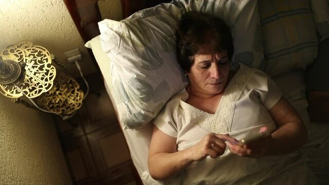 Older woman lying in bed browsing internet on smartphone before sleep