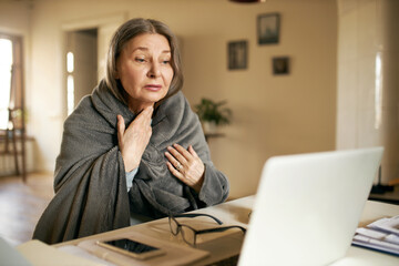 Depressed lonely retired female sitting in front of open portable computer wrapped in plaid,...