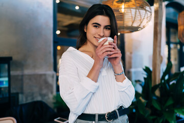 Carefree hipster girl dressed in casual white shirt looking at camera holding disposable cup with caffeine beverage and smiling during rest time in city, happy Caucasian woman enjoying coffee break