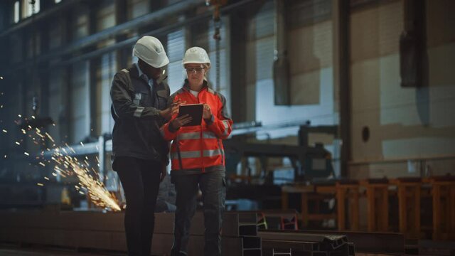 Two Heavy Industry Engineers Walk in Steel Factory, Use Tablet and Discuss Work. Industrial Worker Uses Angle Grinder in the Background. Black African American Specialist Talks to Female Technician.