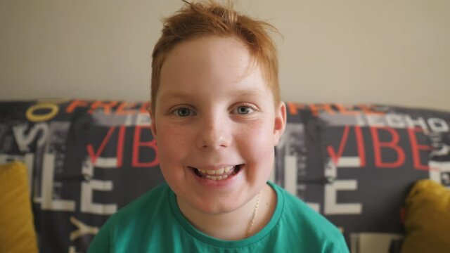 Portrait of happy joyful red-haired boy sitting on the couch and looking into camera. Small male ginger child with freckles laughing indoor. Male face of little smiling kid with positive emotions