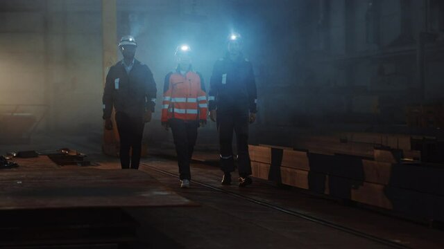 Three Diverse Multicultural Heavy Industry Engineers and Workers Walk in Dark Steel Factory Using Flashlights on Their Hard Hats. Industrial Contractors Walk Towards the Camera Wearing Safety Unofirm.