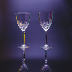 Two crystal wine glasses on long legs stand against a dark blue background. On the edges of the carving, iridescent highlights. Reflection.