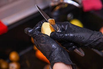 Female hands in black rubber gloves peel potatoes with a knife against the background of a black shell. Cooking. Kitchen