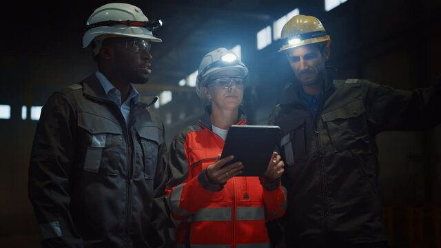 Three Diverse Multicultural Heavy Industry Engineers and Workers in Uniform Stand in Dark Steel Factory Using Flashlights on Their Hard Hats. Female Industrial Contractor is Using a Tablet Computer.