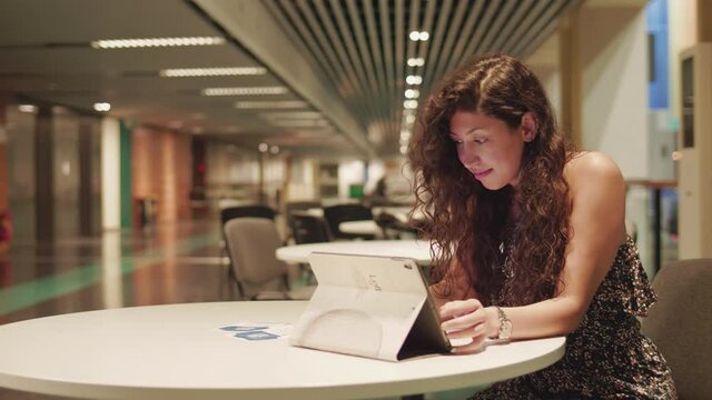Young Pretty Female Sitting by a Table and Checking Online Messages on Tablet.  Urban Girl in Modern Indoor Environment, Static Full Frame