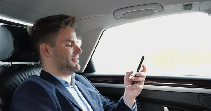 Close Up of a Relaxed Calm Confident Businessman is texting Messages while riding in Luxury Car. A handsome Man is thoughtfully riding News on the Smartphone in his Elegant Car with his own Driver.