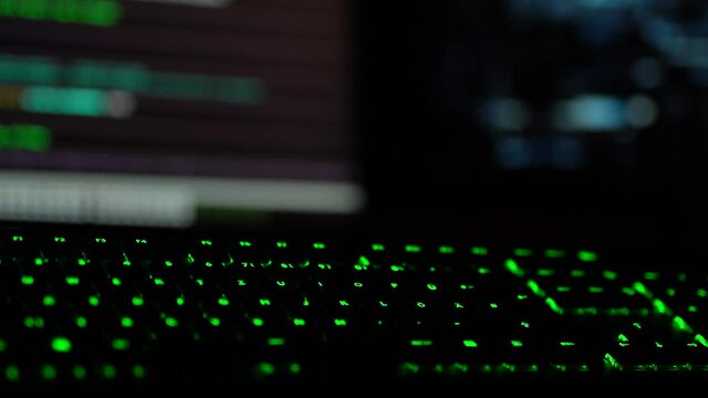 Cybersecurity concept. Closeup of green lit keyboard in dark room with hacker code blinking on screen in the background
