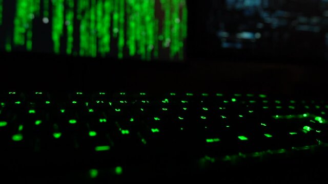 Cybersecurity concept. Slowmotion focus shift of green lit keyboard in dark room with hacker code blinking on screen in the background