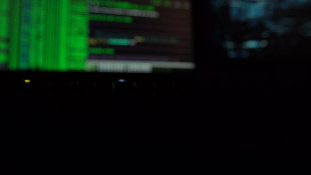 Cybersecurity concept. Closeup of green blinking keyboard in dark room with hacker code on screen in the background