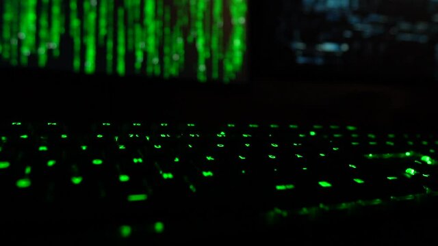Cybersecurity concept. Slowmotion closeup of green lit keyboard in dark room with hacker code blinking on screen in the background