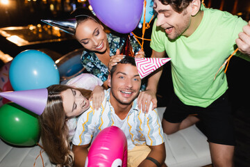 Selective focus of friends hugging young man in party cap near balloons and swimming pool at night