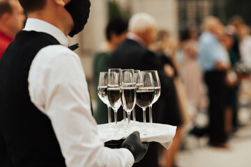 Waiter in black mask and gloves hold tray with glasses of champagne. Waiter holding a serving tray full of drinks in champagne glasses. Wearing masks during coronavirus.