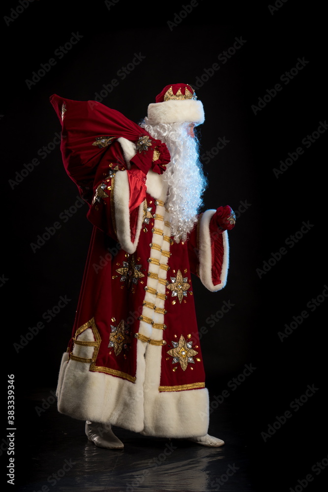 Wall mural Santa Claus in a red fur coat, with a white beard and a bag of gifts posing on a black background