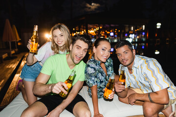 Selective focus of friends with beer bottles looking at camera outdoors at night