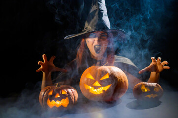 An ominous witch in a hat conjures over a jack-o-lantern. Traditional halloween characters. Mystical fog creeps over pumpkins with carved terrible faces. An evil witch casts a spell.
