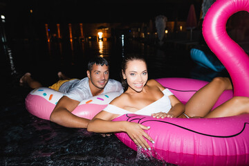 Selective focus of young couple looking at camera while swimming on rings in pool at night