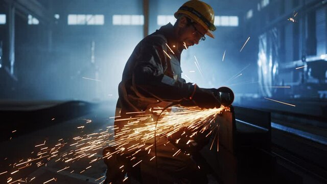 Heavy Industry Engineering Factory Interior with Industrial Worker Using Angle Grinder and Cutting a Metal Tube. Contractor in Safety Uniform and Hard Hat Manufacturing Metal Structures.