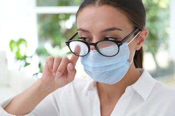 Woman wiping foggy glasses caused by wearing medical mask indoors, closeup